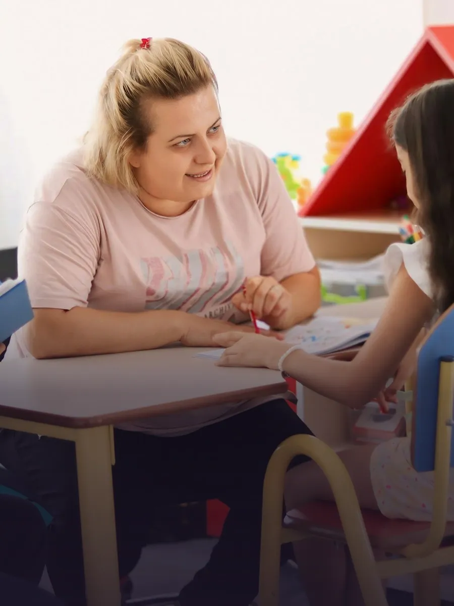 Teacher talking with a parent at a school desk, a pupil beside her